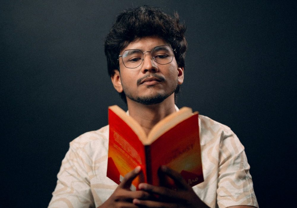 A man sitting reading an open book of poetry to an audience.