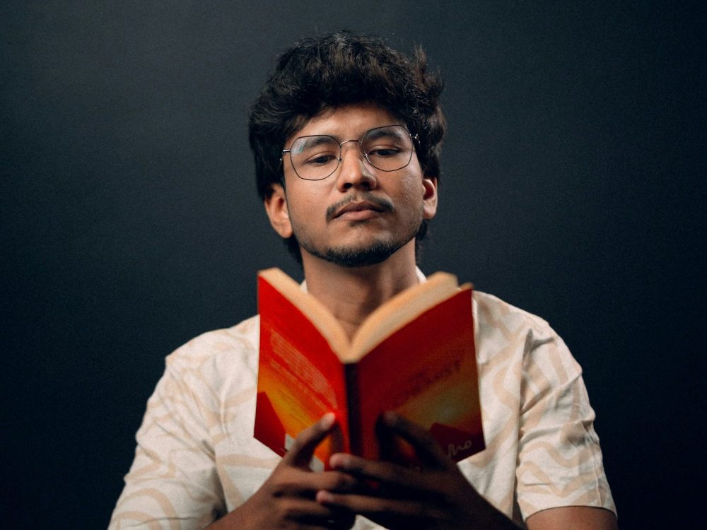 A man sitting reading an open book of poetry to an audience.