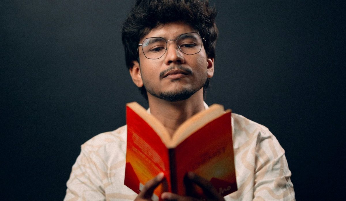 A man sitting reading an open book of poetry to an audience.
