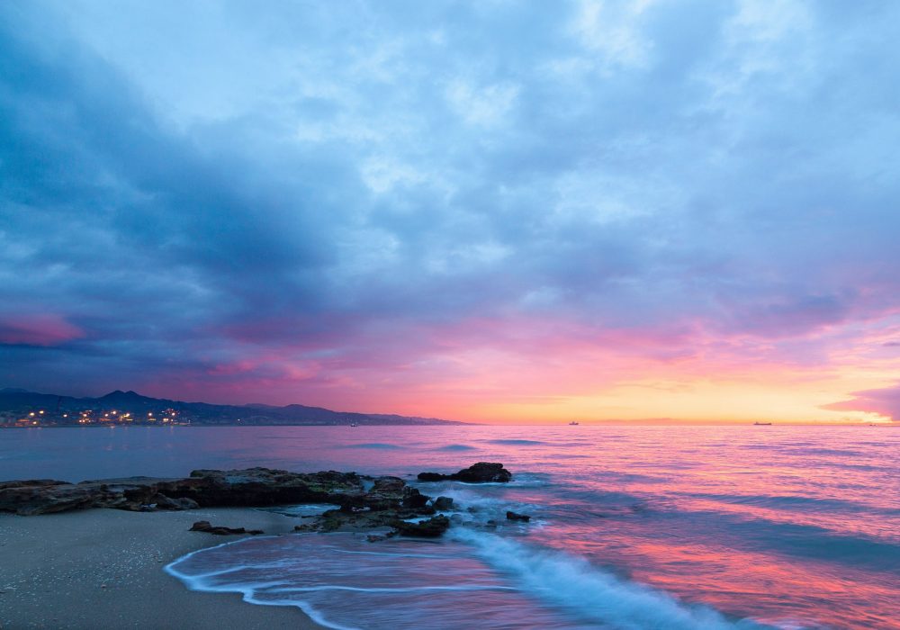 Rocky beach at sunset with pink and blue sky reflecting on calm sea.