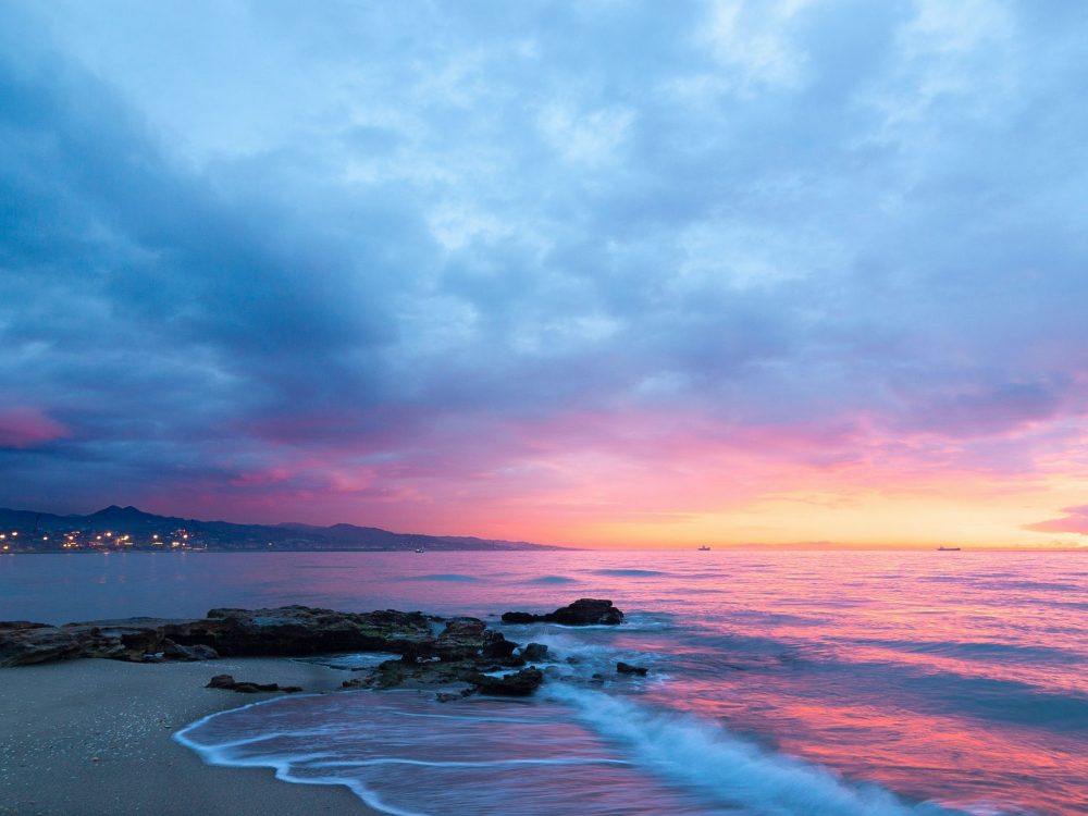Rocky beach at sunset with pink and blue sky reflecting on calm sea.