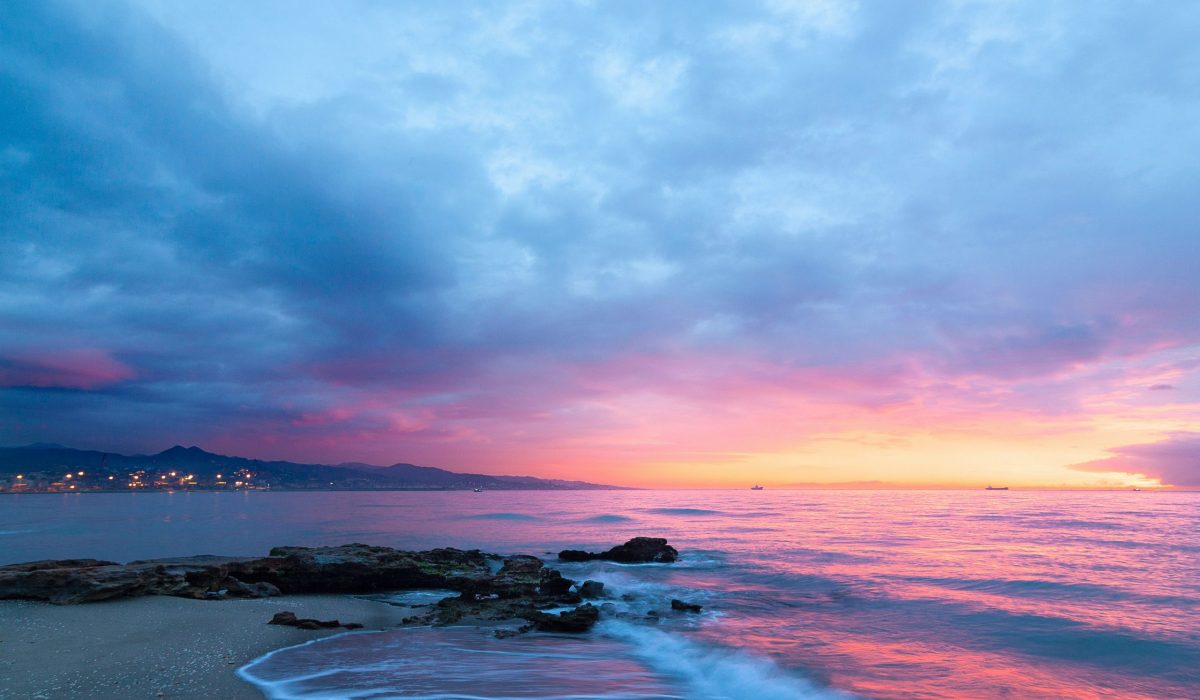 Rocky beach at sunset with pink and blue sky reflecting on calm sea.